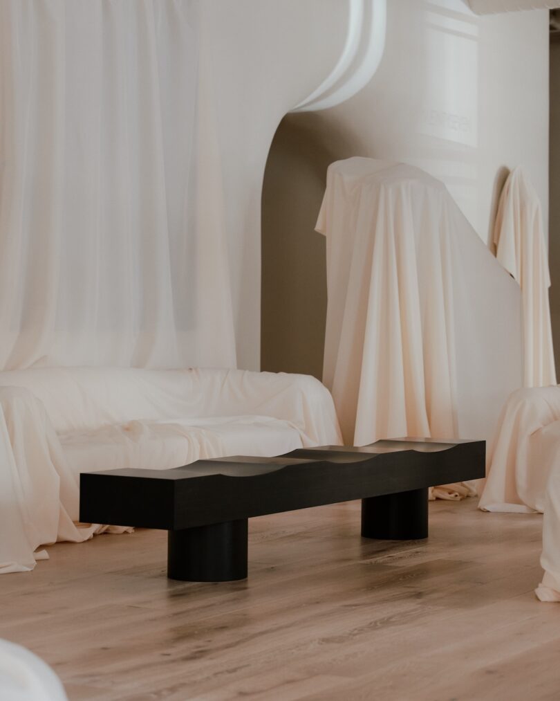 A modern black table stands on a wooden floor, surrounded by furniture covered in white sheets, in a minimalist, neutral-toned room.