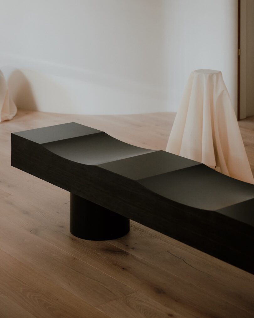 A modern black bench with a wavy surface stands on a light wooden floor, with draped cloth-covered objects in the background.