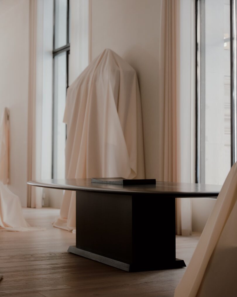 Minimalist room with large windows, having beige curtains. A dark modern table with a book placed on it is situated near a covered object in the background. Light wooden flooring is evident.