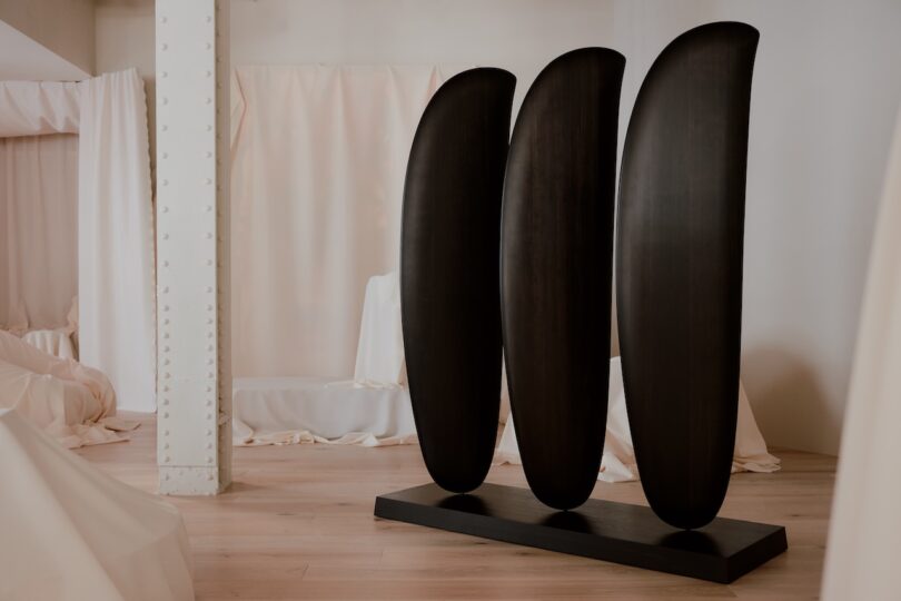 A dark wooden sculpture with three vertical, curved panels mounted on a rectangular base placed in a room with white drapes and light wood flooring.