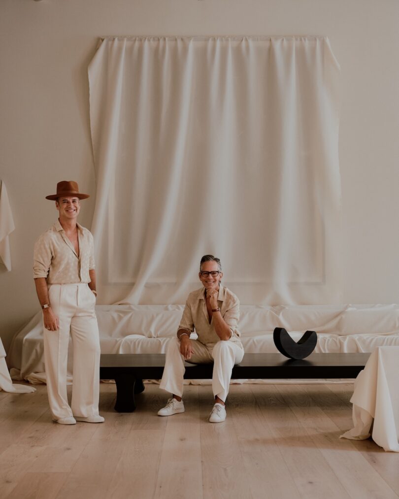 Two individuals in light-colored clothing pose in a minimalistic room with white draped fabric. One stands wearing a hat, and the other sits on a bench with a black geometric sculpture.