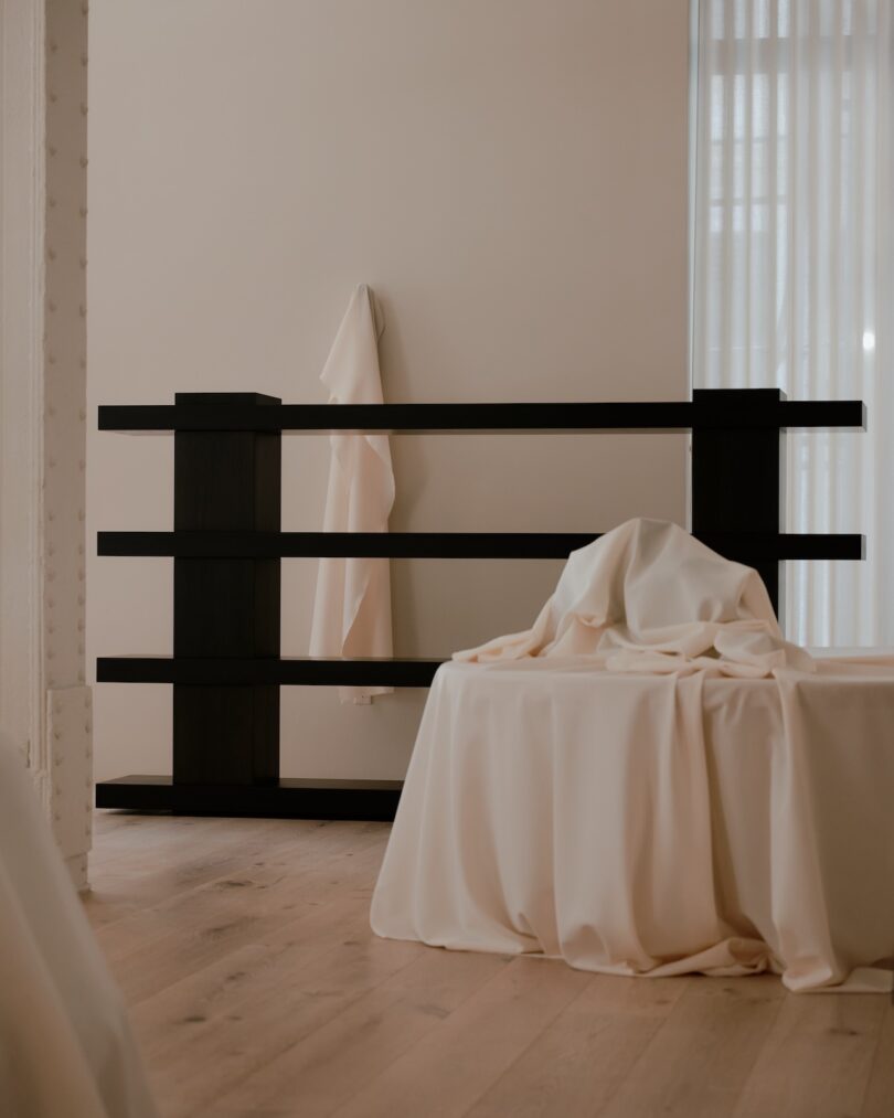 A minimalist room with a black wooden shelving unit, a light-colored cloth hanging on it, and a covered object on a low, cloth-covered table. Light wooden floor and sheer curtains in the background.