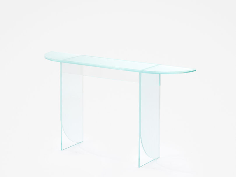 A transparent glass table with a rectangular surface and two curved end panels serving as legs, set against a white background