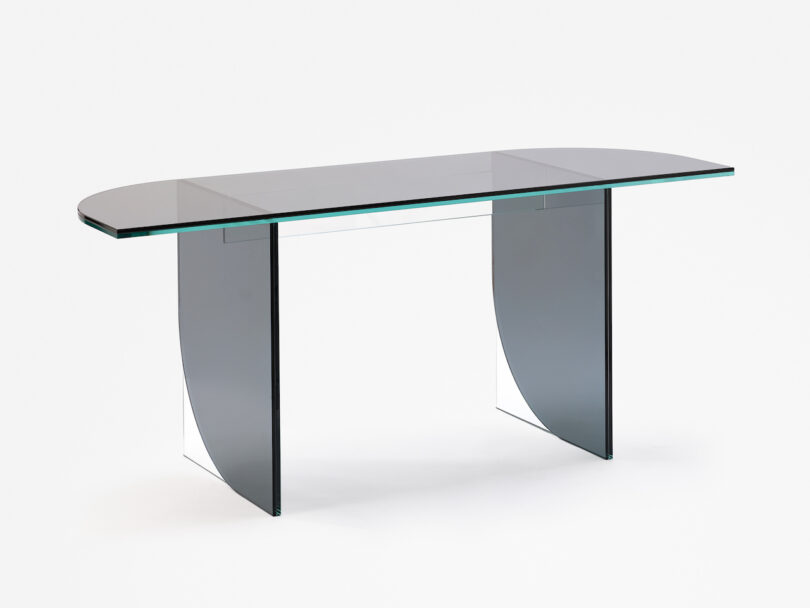 A glass-top rectangular table with two curved black base supports is displayed against a white background