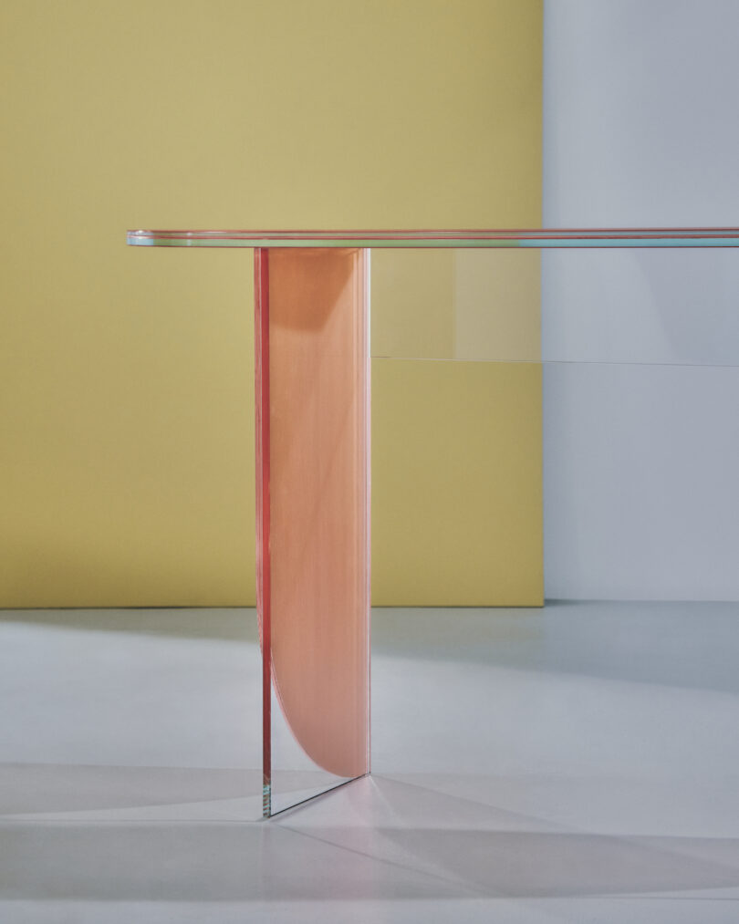 A glass table with a modern design, featuring a clear glass top and a distinctive, curved, pinkish-orange support leg, set against a yellow and white background