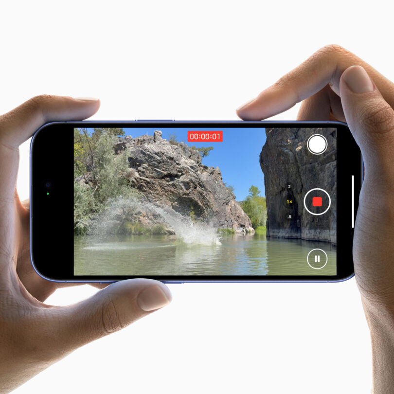 Hands holding a smartphone recording a splash in a river with rocky cliffs and trees in the background. The screen displays a red recording timer and icons for recording control.