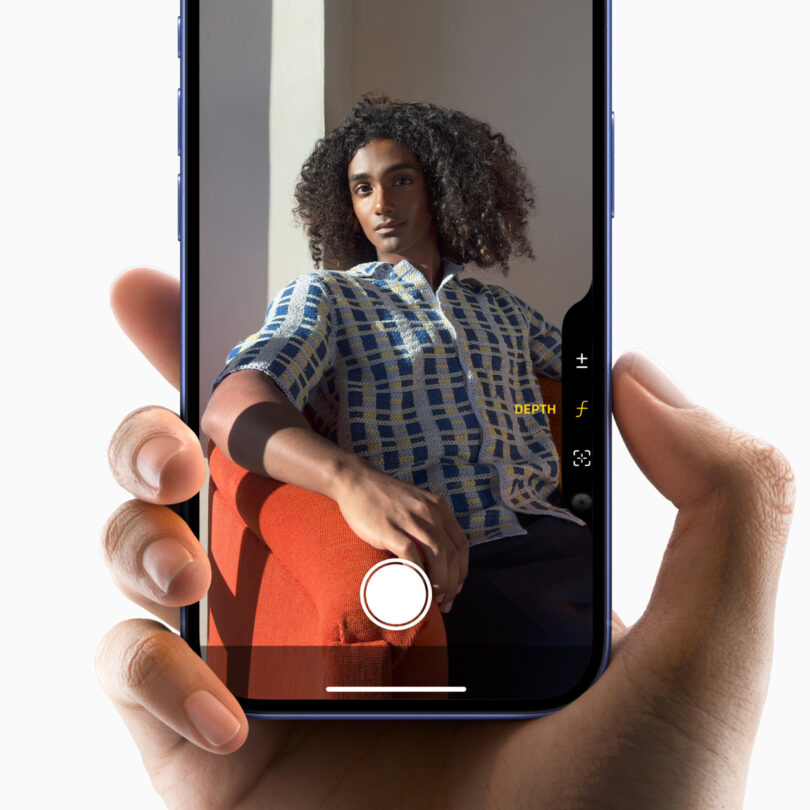 A hand holding a smartphone displaying a photo of a person with curly hair sitting on a red chair, wearing a blue and white checkered shirt. The phone camera interface is visible on the screen.