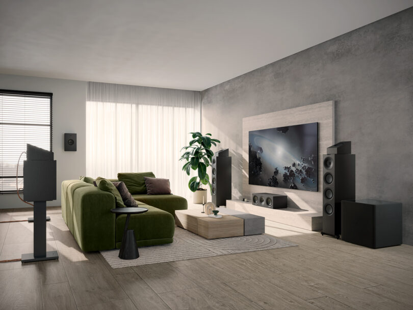 A modern living room featuring a green sectional sofa, a large flat-screen TV mounted on a gray wall, surround sound speakers, a wooden coffee table, and a tall indoor plant.