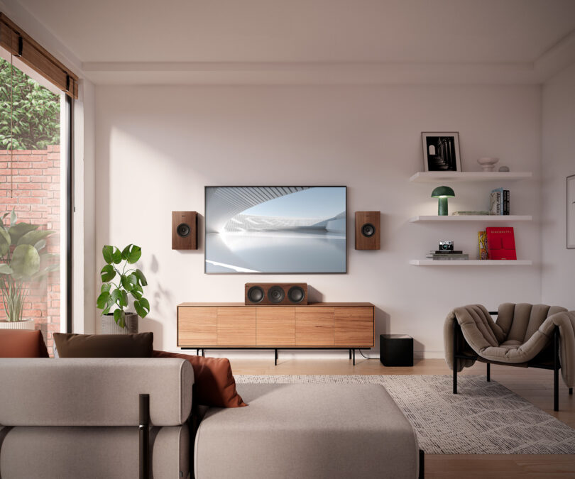 Modern living room with wall-mounted TV, wooden console, and shelves. Includes sofa, armchair, speakers, plants, and minimalist decor. Sunlight enters from a large window on the left.