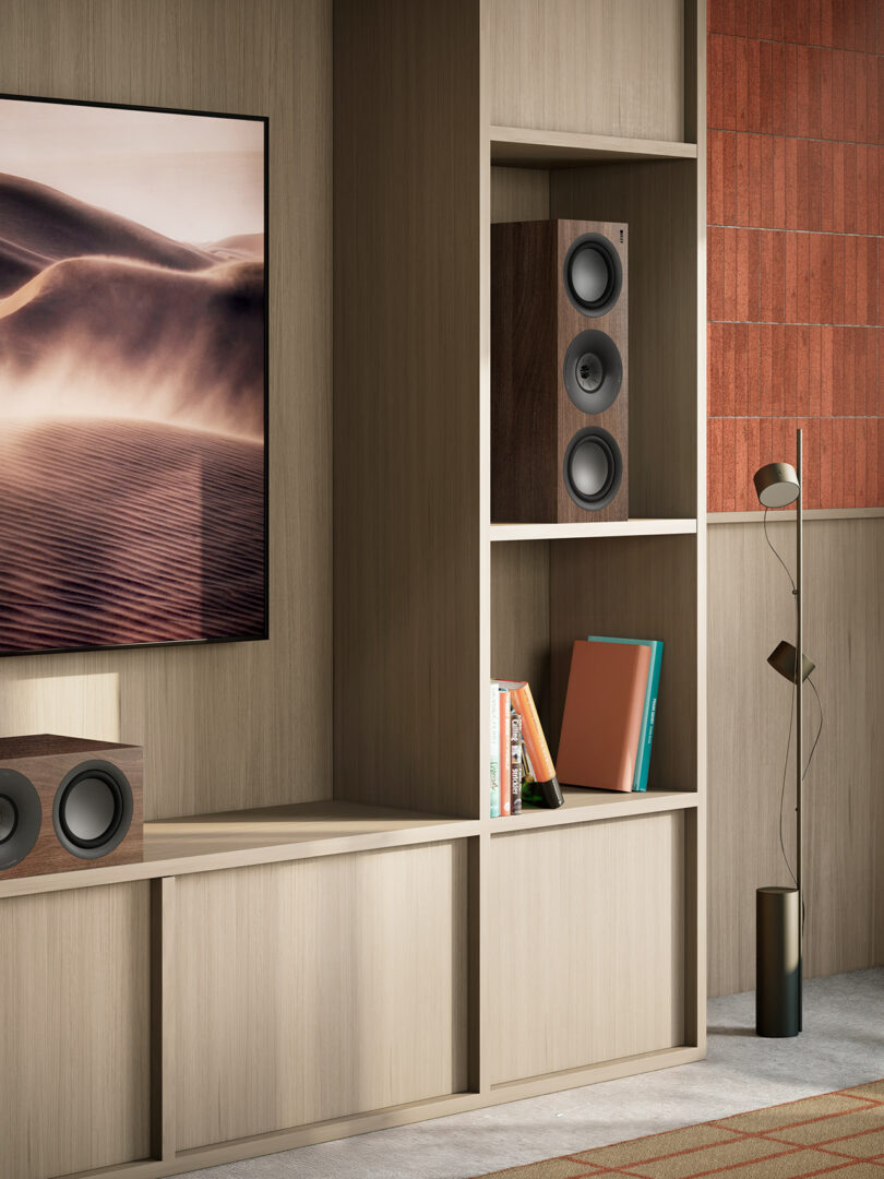 Modern living room with beige wooden furniture, a landscape photo on the wall, speakers, books, and a floor lamp with multiple small shades.