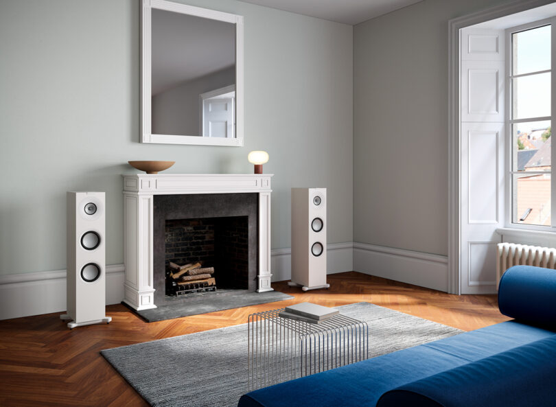 A modern living room with a fireplace, two floor-standing speakers, a mirror above the mantle, a small lamp, a blue sofa, a glass coffee table, and a large window on the right side.