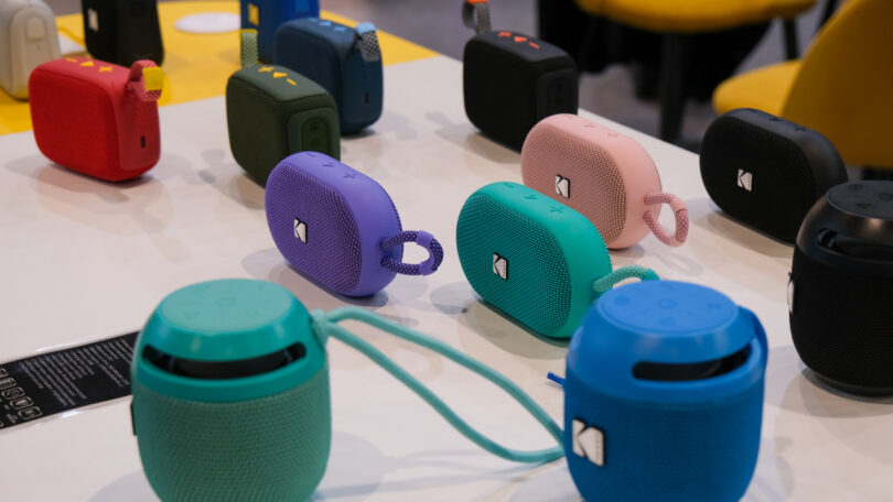 Various colorful portable Bluetooth speakers are displayed on a table, featuring different shapes and sizes, with some having fabric straps attached.