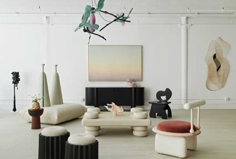 A modern art gallery features a variety of abstract furniture and sculptures in a minimalist setting, including a central cushioned coffee table, unique chairs, and wall art