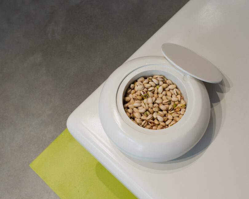 A white ceramic bowl with a lid holds a bunch of pistachio nuts, placed on a white and green surface