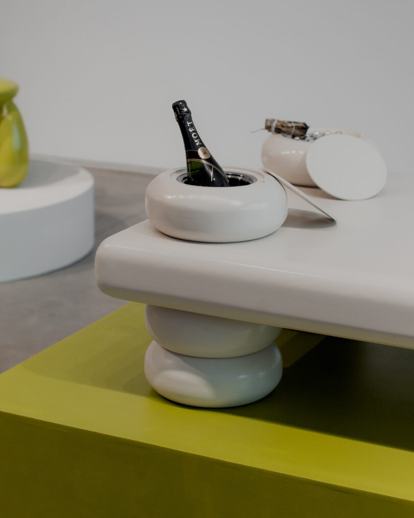 A modern white table with chunky legs holds a white bowl with a bottle of sparkling wine and a separate white dish nearby. Another white furniture piece and a green object are in the background