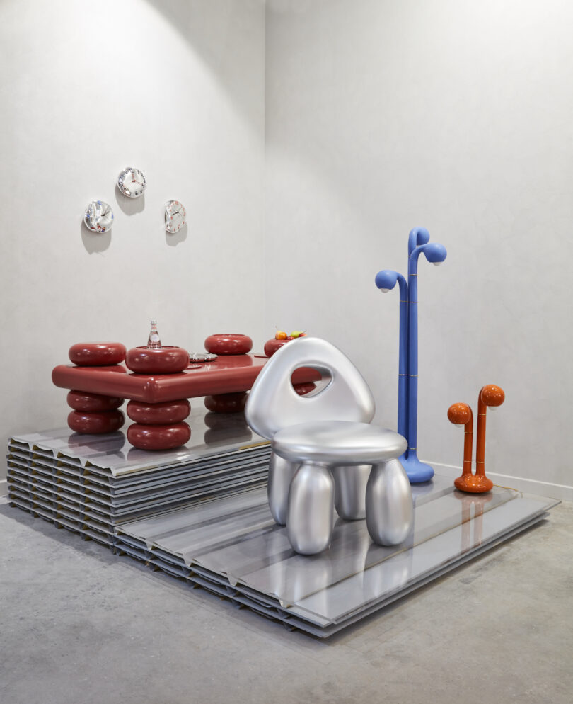 A futuristic room setup featuring a silver metallic chair, a red cushioned table set, and abstract sculptures in blue and orange on a raised platform with a minimalist wall backdrop