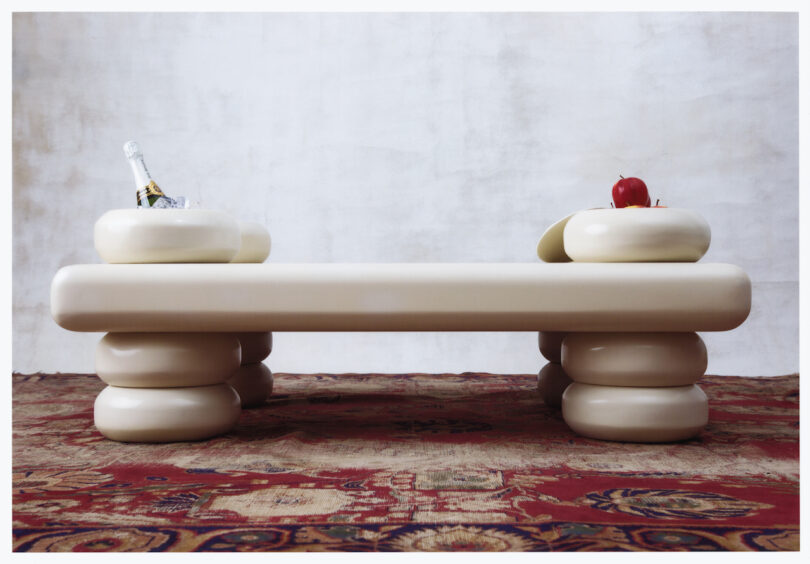 A modern beige table with rounded legs on a patterned red carpet. The table has built-in oval bowls one holding an ice bucket with a bottle and another holding fruit including an apple