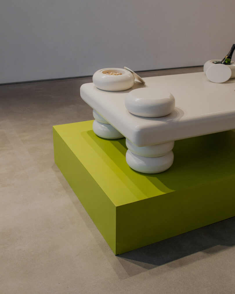 A white, rectangular, ceramic table with rounded ends and circular supports, displayed on a lime green platform in a minimalist setting