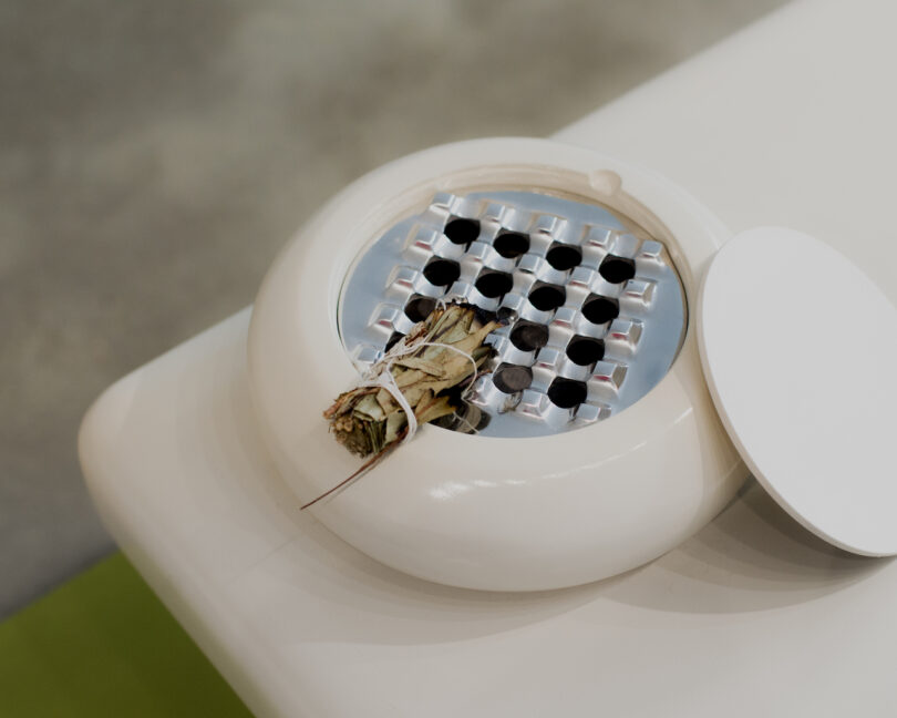 A smudge stick of herbs rests on a round, white ceramic incense holder with a metal grill, positioned on a white surface