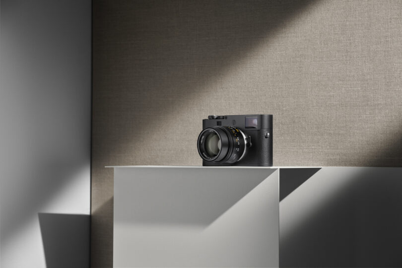 A black camera with a large lens is placed on a minimalist, grey pedestal under a spotlight against a textured, neutral-colored background.