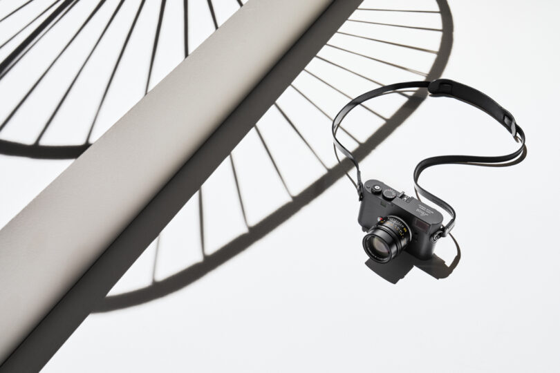 A camera with a strap lies on a white surface with the shadow of a bicycle wheel cast across the background.