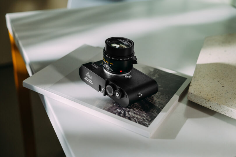 A black Leica camera rests on a white table, atop a partially visible book with a blurred image on its cover. The scene is softly lit with natural light.