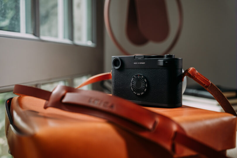 A black camera with a round dial sits on an orange leather bag near a window.