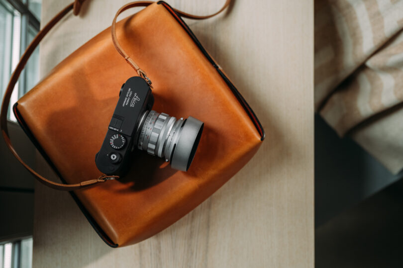 A camera with a strap rests on a tan leather bag placed on a light-colored wooden surface.