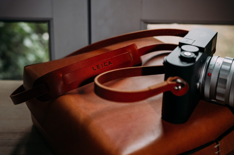 Close-up of a Leica camera with a brown leather strap and a brown leather bag in the background.