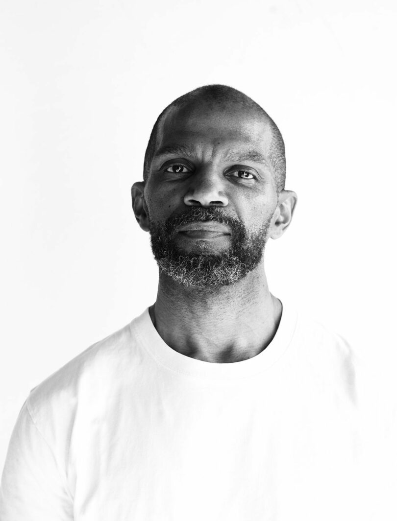 A man with a short beard and a shaved head wears a plain white shirt and looks directly at the camera. The image is in black and white