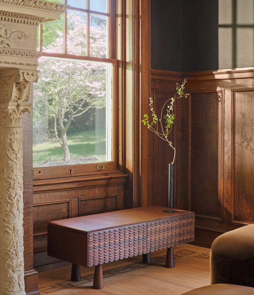 A wooden room corner with a window, a detailed carved fireplace, a small wooden bench, and a tall slender vase holding a branch with leaves and small white flowers. A tree is visible outside