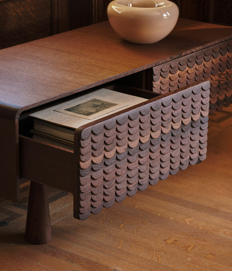 A wooden side table with a drawer partially open, displaying stacked books. The drawer features a decorative, scalloped pattern, and a ceramic bowl sits on top of the table