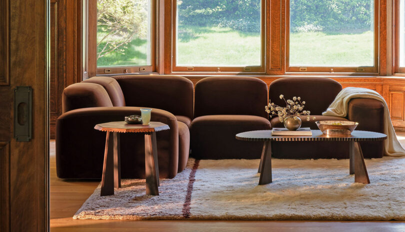 A living room with a large brown sectional sofa, two black coffee tables, a small side table, and a cream-colored rug. Sunlight streams in through the large windows in the background