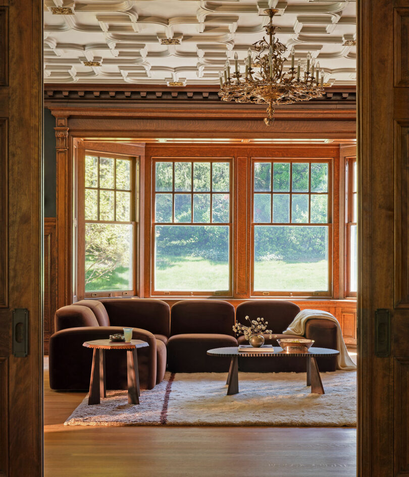 A living room features a dark brown sofa, two circular tables, a light rug, and large windows offering a view of greenery. The room has wooden paneling, an ornate ceiling, and an elaborate chandelier