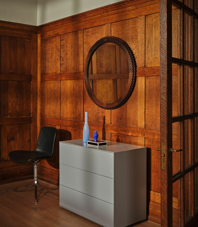 A modern grey chest of drawers with two blue bottles and a book on top stands against a paneled wooden wall, with a round mirror above and a black chair to the left