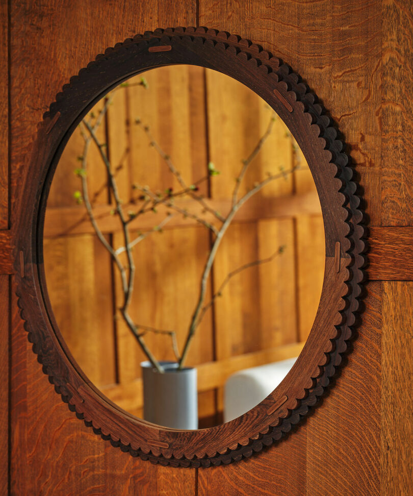 An oval mirror with a dark, scalloped wooden frame reflects a vase with branches against a wooden paneled background