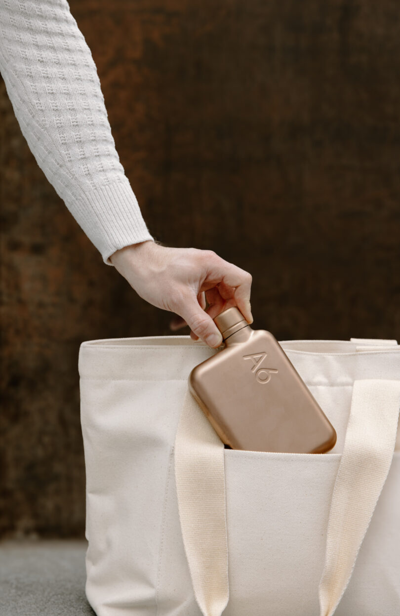 A person in a white knit sweater places a bronze flask into the front pocket of a beige tote bag