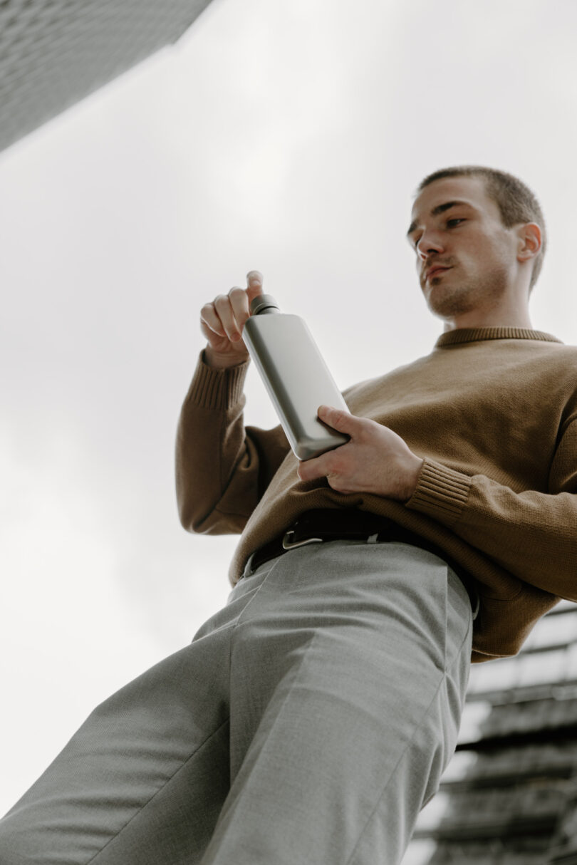 Man in a brown sweater and gray pants holding a water bottle, looking down with buildings in the background