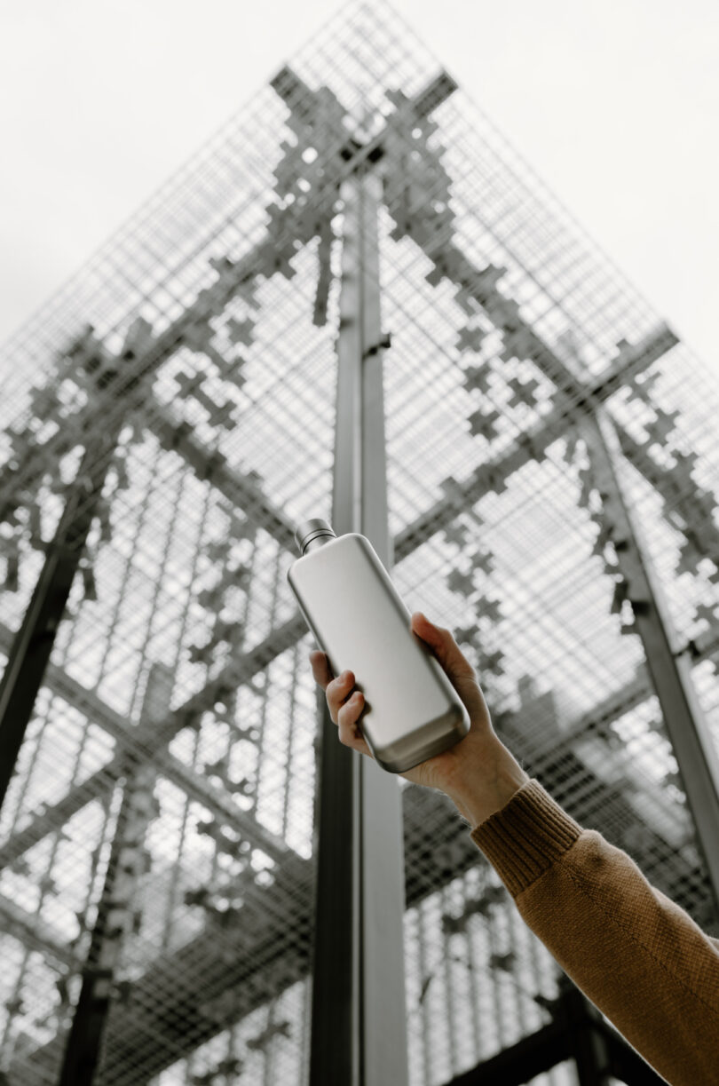 A hand in a brown sleeve holds up a silver bottle in front of a tall, metal lattice structure