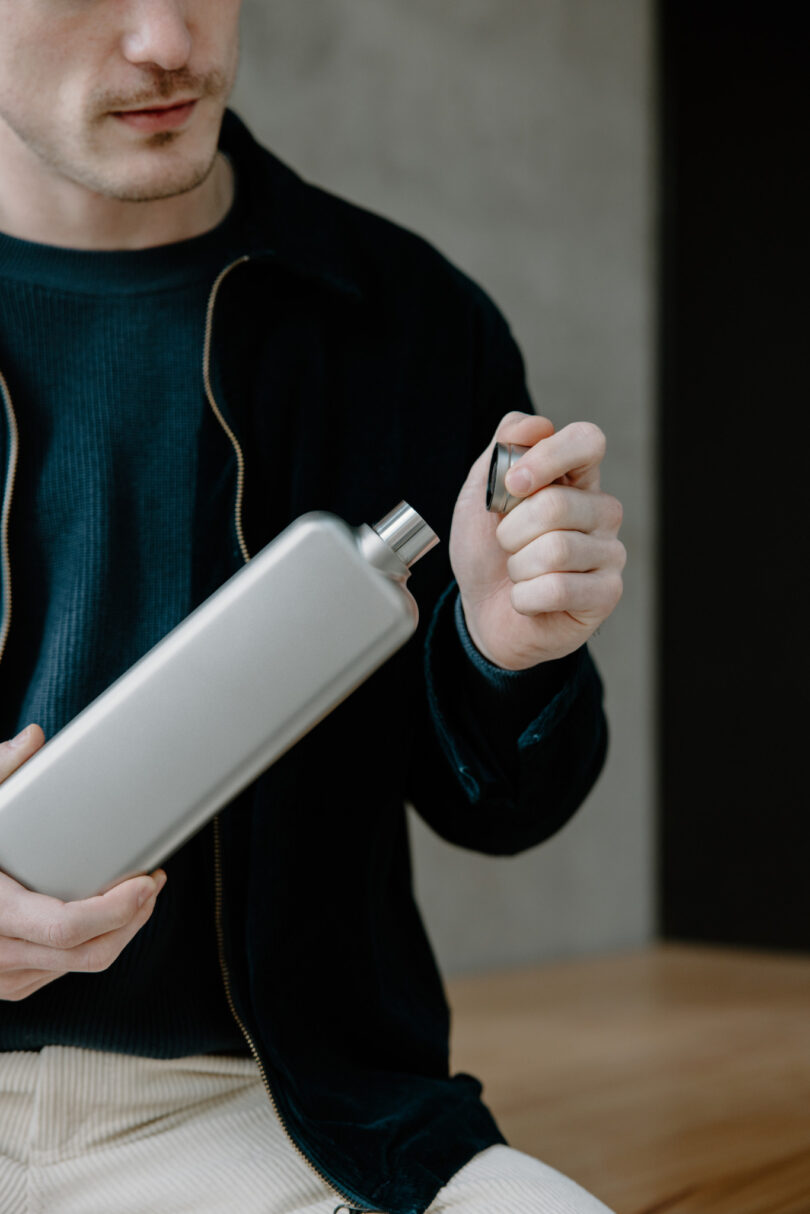 Person in a dark jacket opening a rectangular silver bottle indoors