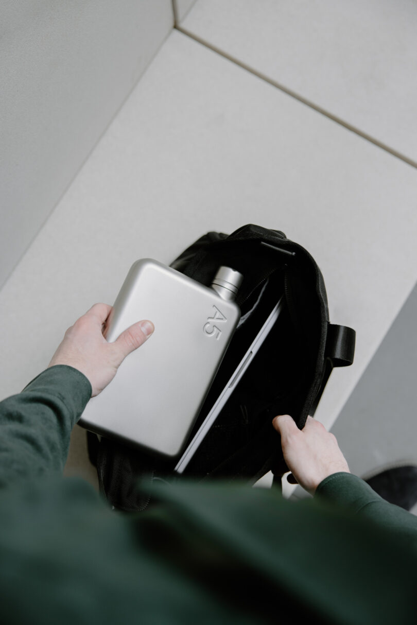 A person places a sleek, silver A5-sized water bottle into an open black bag