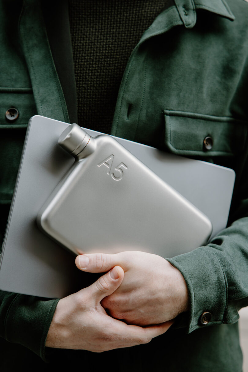 A person in a green jacket holds a silver A5 flask and a closed laptop