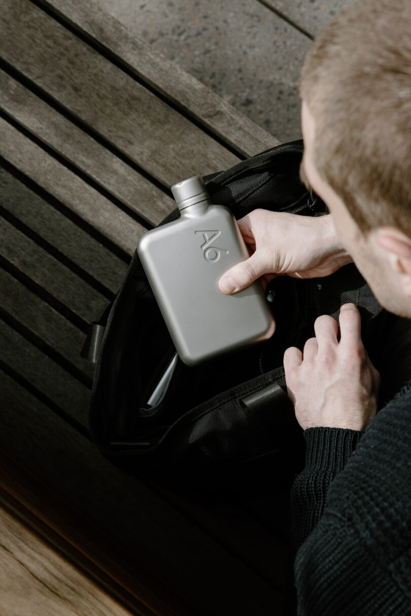 A person is placing a metallic flask into a black bag while sitting on a wooden bench