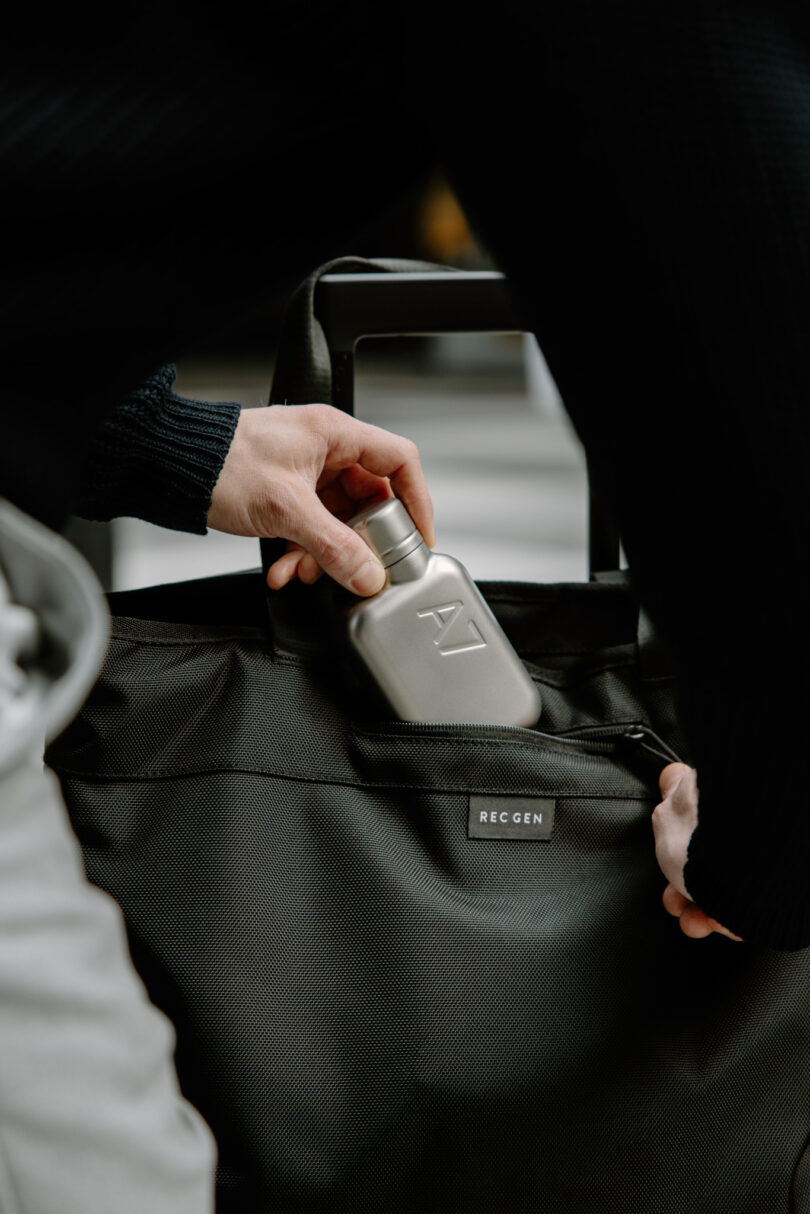 Close-up of a person placing a small silver bottle into the pocket of a black RECCEN bag