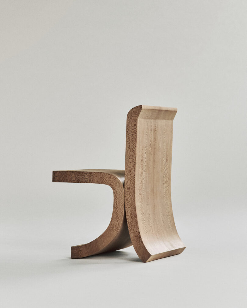 A modern, minimalist wooden chair with smooth, curved lines and a sculptural design against a plain, light grey background.