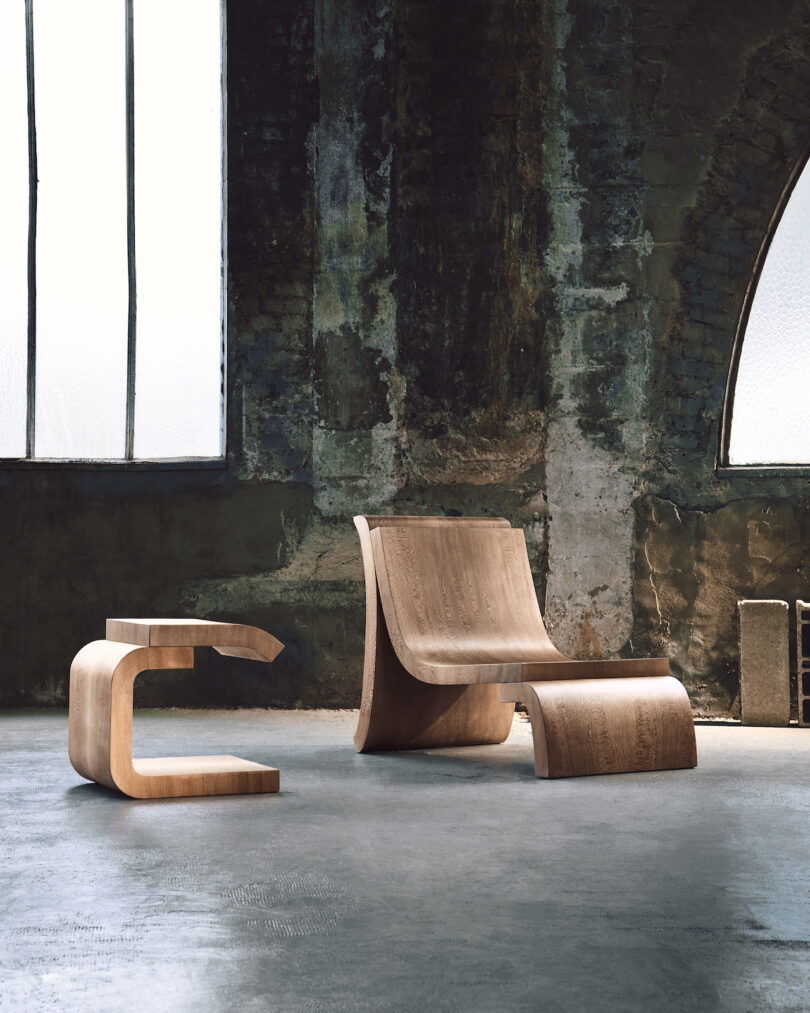 A modern wooden chair with an unconventional, curvy design is paired with a matching side table in a rustic room with large windows and distressed walls.