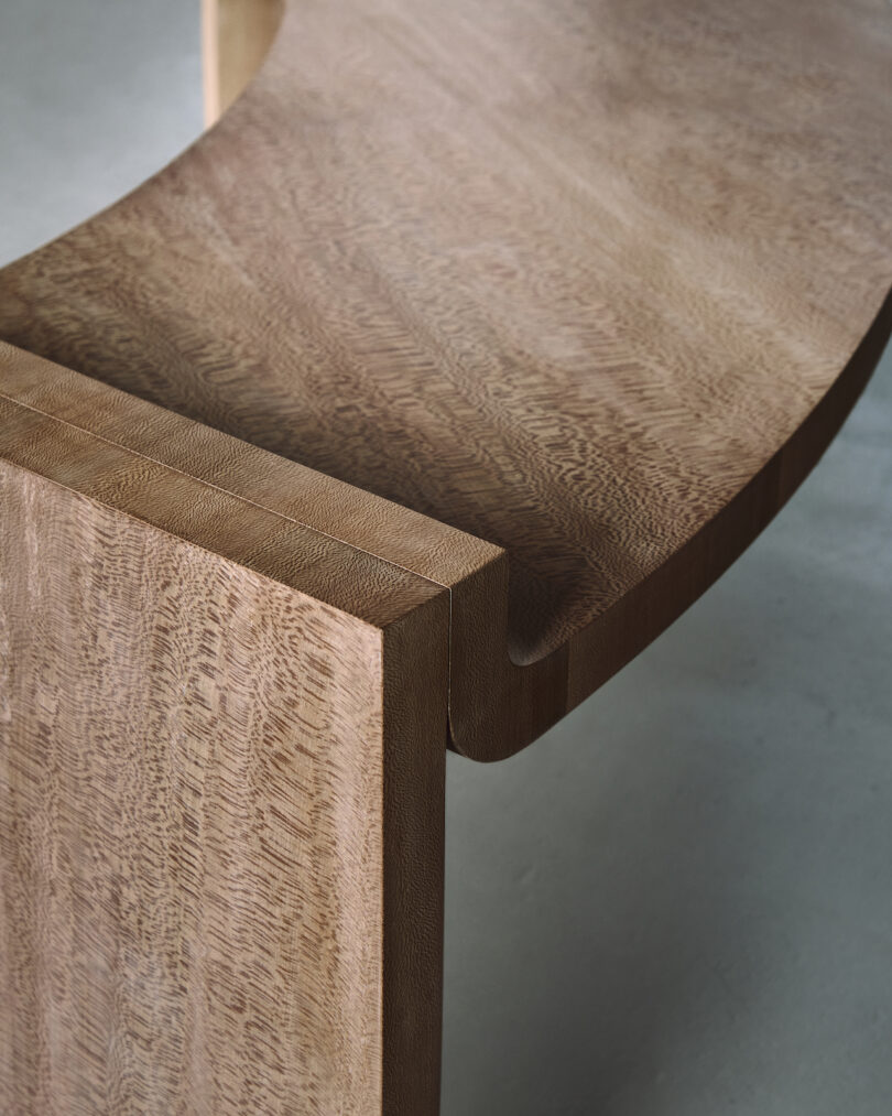 Close-up of a curved wooden bench with a smooth grain texture, showcasing the craftsmanship and joinery details against a neutral background.