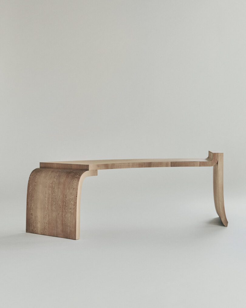 A modern, minimalist wooden bench with a curved base on one side and a single leg on the other, set against a plain background.