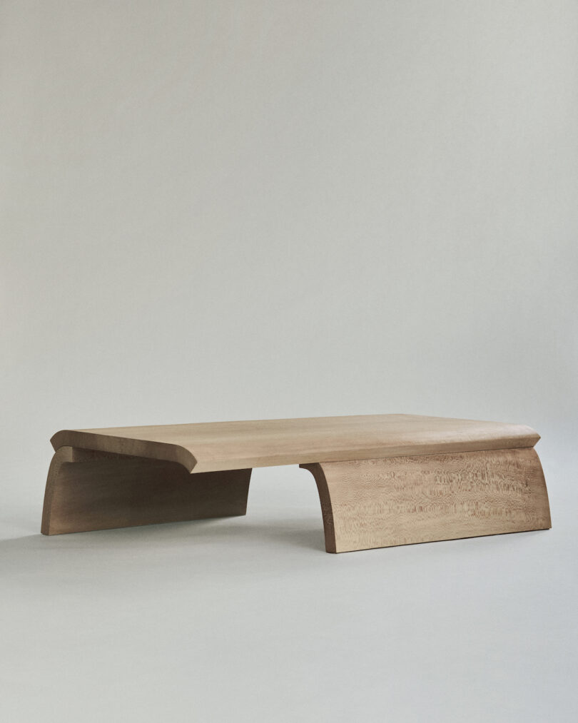 A wooden coffee table with a minimalist design featuring a rectangular surface and curved legs. The table has a natural finish and smooth edges.