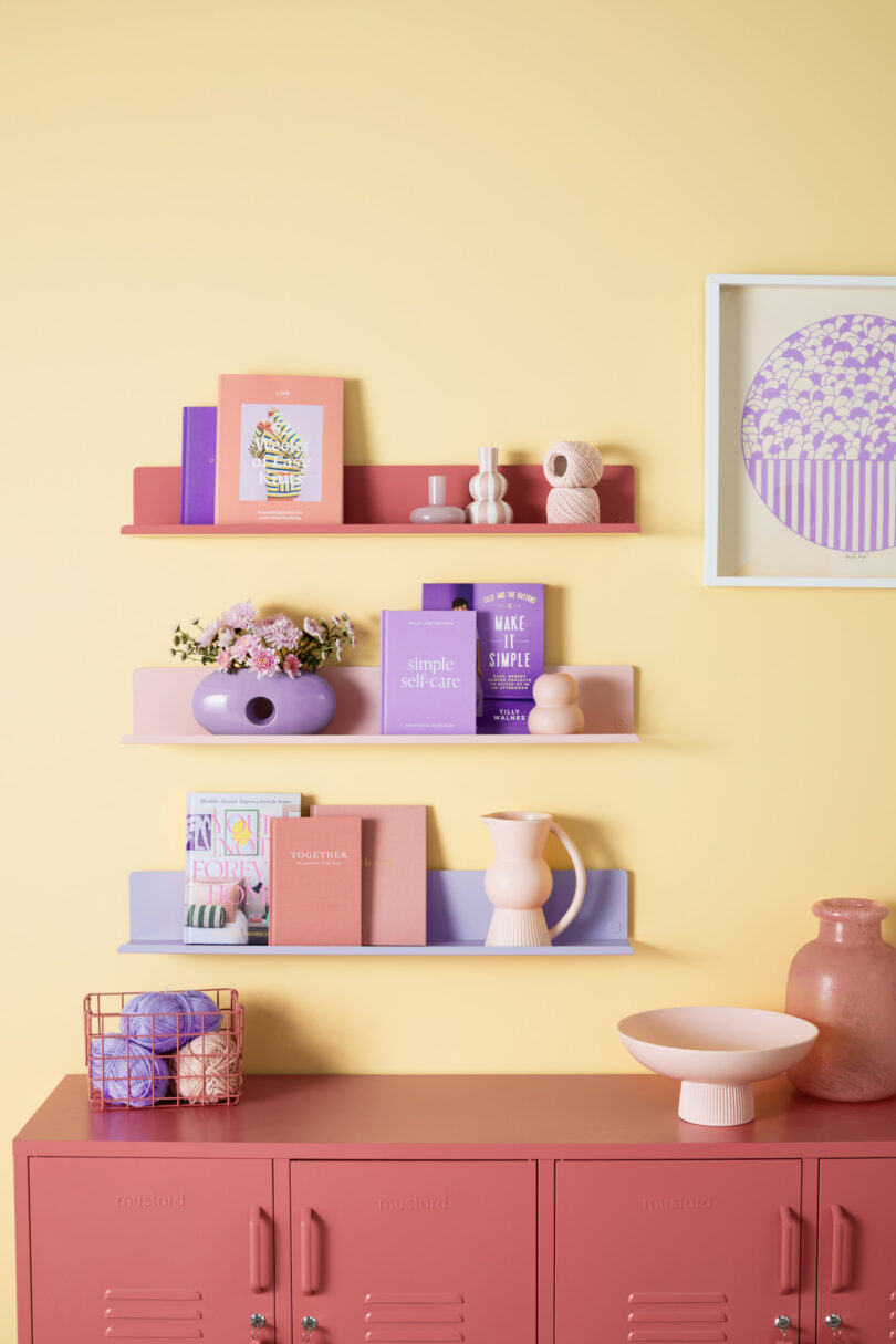 A pastel-themed room with colorful shelves displaying books, flowers, and vases on a yellow wall, above a pink storage cabinet with various decorative items